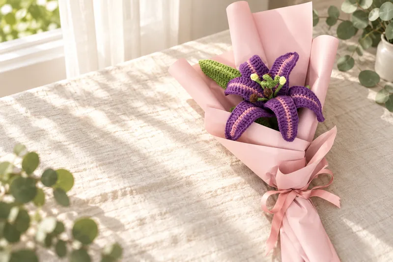 A crocheted purple lily with pink stripes in pink paper wrap, on a linen cloth in the morning sun