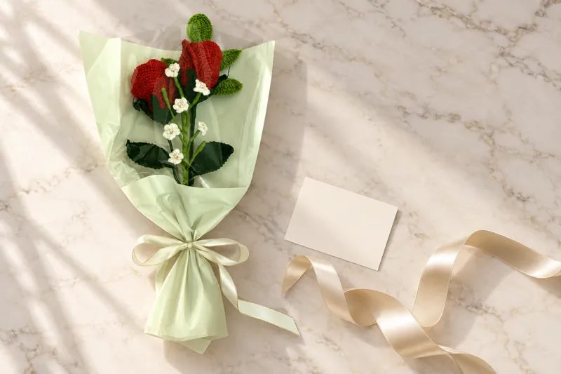Two crocheted red roses with small white blossoms in pale green wrap on a marble table, with a ribbon and a blank card